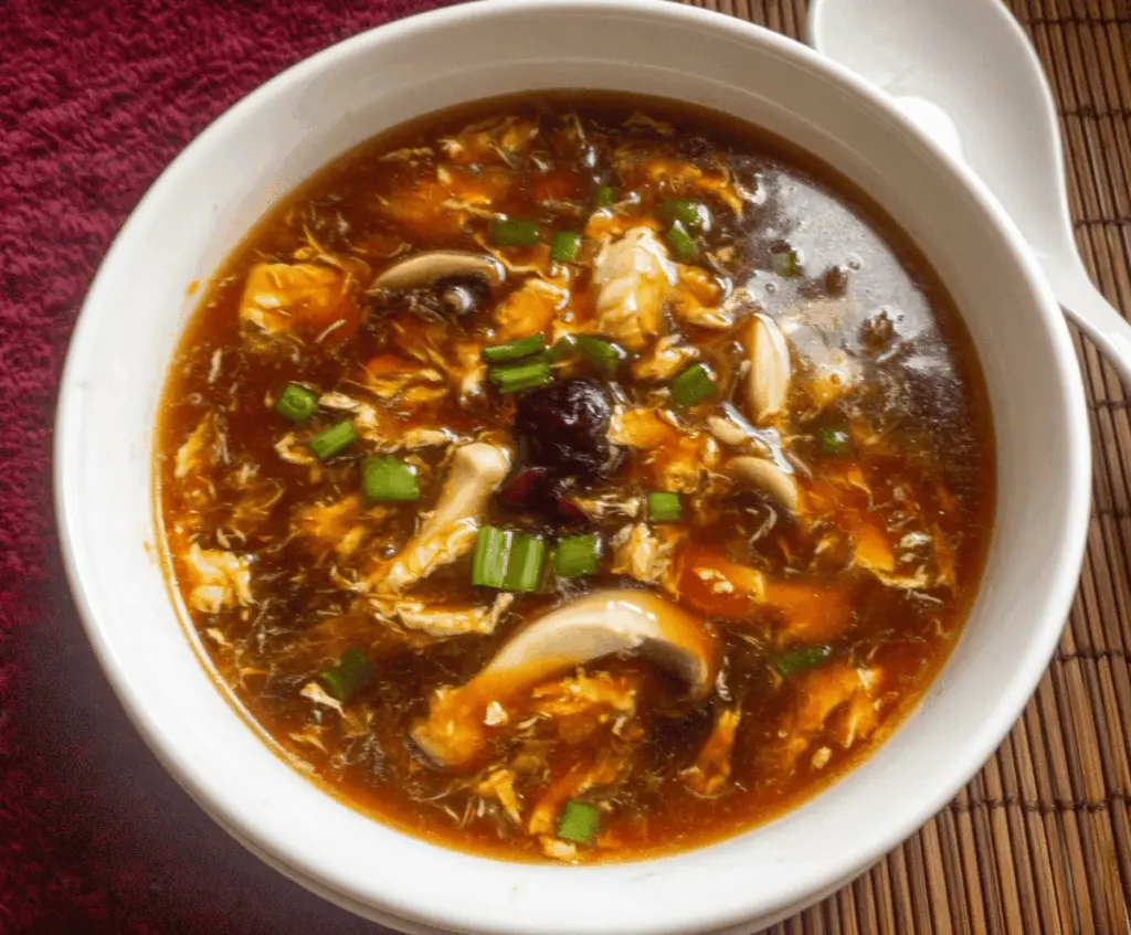 A steaming bowl of Chinese Hot and Sour Soup garnished with green onions, tofu, mushrooms, and sliced bamboo shoots, showcasing a flavorful, spicy, and tangy Asian appetizer.
