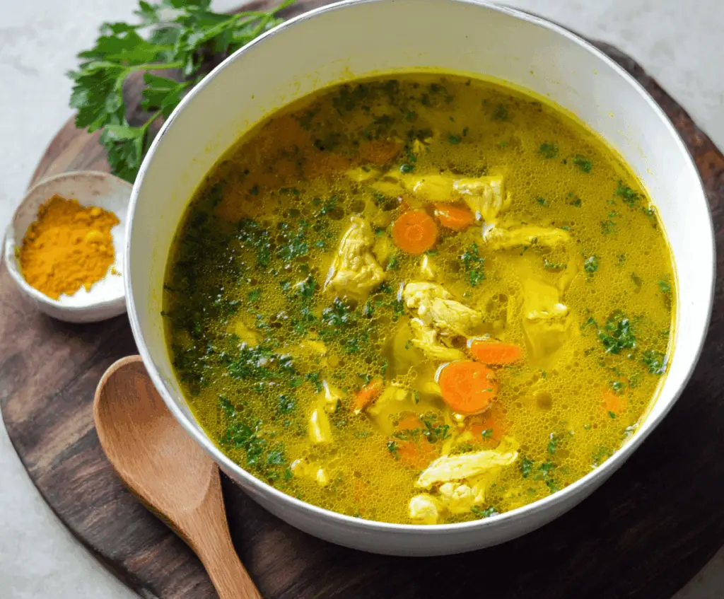 A bowl of anti-inflammatory turmeric chicken soup garnished with fresh herbs and spices, showcasing vibrant yellow turmeric broth with tender chicken pieces and vegetables.