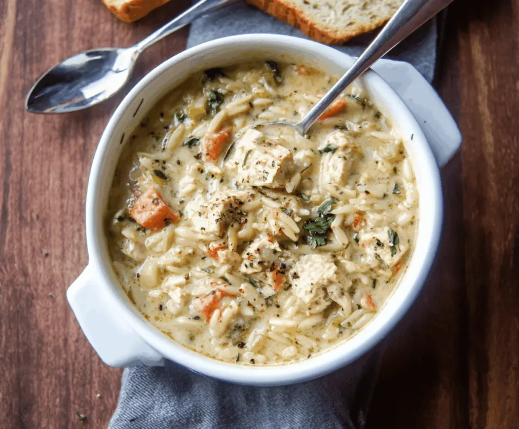 A steaming bowl of creamy chicken orzo soup garnished with fresh herbs and served with bread on the side.